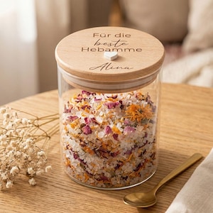 Könnte beinhalten: Ein durchsichtiges Glasgefäß gefüllt mit einer Mischung aus weißem Badesalz, getrockneten rosa Rosenblättern und orangefarbenen Ringelblumen. Der runde Holzdeckel ist mit dem Text "Für die beste Hebamme" und "Alina" sowie einem kleinen weißen Herz graviert.