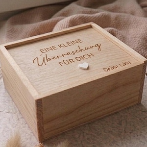 Könnte beinhalten: Eine rechteckige, helle Holzschachtel mit Schiebedeckel. Der Deckel ist mit dem deutschen Text "EINE KLEINE ÜBERRASCHUNG FÜR DICH" und "Deine Lina" graviert und mit einem kleinen, weißen, herzförmigen Schmuckelement unter dem Text versehen.