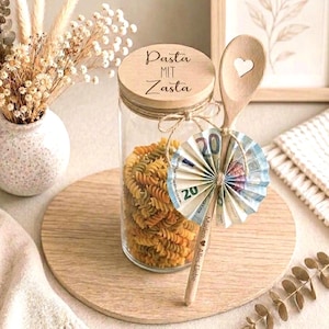 Op de afbeelding: Een doorzichtige glazen pot gevuld met rotini pasta, met een houten deksel gegraveerd met de tekst Pasta MIT Zasta. Een houten lepel met een hartvormige uitsparing is met touw aan de pot bevestigd, versierd met een waaiervormig gevouwen bankbiljet.