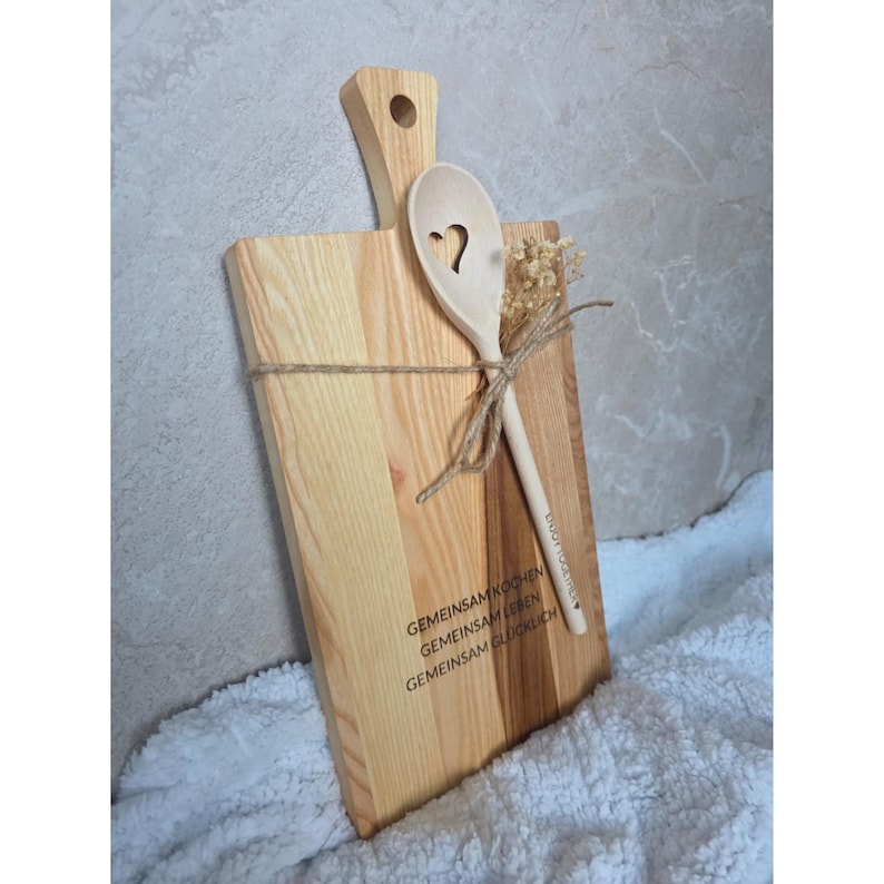 May include: A rectangular light-colored wooden cutting board with a handle and a hole, featuring the engraved text GEMEINSAM KOCHEN GEMEINSAM LEBEN GEMEINSAM GL&Uuml;CKLICH. A wooden spoon with a heart-shaped cutout and dried flowers are tied to the board with twine.