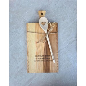 May include: A rectangular wooden cutting board with a handle and a wooden spoon tied together with twine. The board features the text GEMEINSAM KOCHEN GEMEINSAM LEBEN GEMEINSAM GL&Uuml;CKLICH. The spoon has a heart cutout and the text ENJOY TOGETHER.