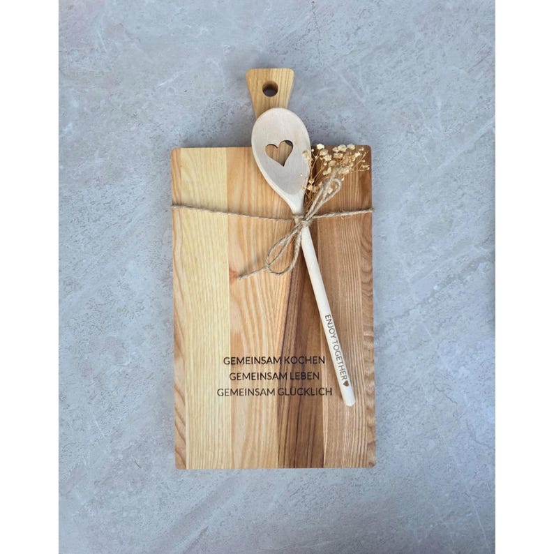 May include: A rectangular wooden cutting board with a handle and a wooden spoon tied together with twine. The board features the text GEMEINSAM KOCHEN GEMEINSAM LEBEN GEMEINSAM GL&Uuml;CKLICH. The spoon has a heart cutout and the text ENJOY TOGETHER.