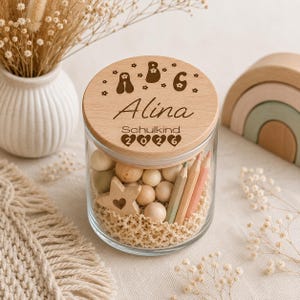 May include: A clear glass jar with a round wooden lid engraved with the letters A, B, C, the name Alina, the word Schulkind, and the year 2026. The jar is filled with small wooden spheres, a wooden star, and colored pencils on a textured base.
