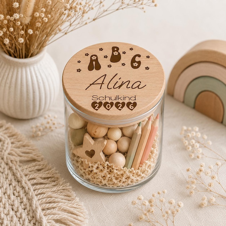 May include: A clear glass jar with a round wooden lid engraved with the letters A, B, C, the name Alina, the word Schulkind, and the year 2026. The jar is filled with small wooden spheres, a wooden star, and colored pencils on a textured base.