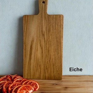 May include: A rectangular wooden cutting board with a handle and a hole for hanging. The board features a visible wood grain pattern and is positioned upright. The word Eiche is printed in black text to the right of the board.