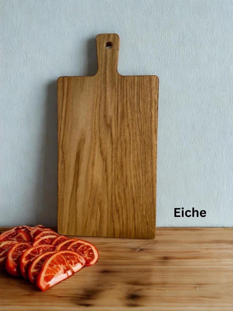May include: A rectangular wooden cutting board with a handle and a hole for hanging. The board features a visible wood grain pattern and is positioned upright. The word Eiche is printed in black text to the right of the board.