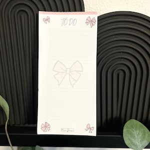 Könnte beinhalten: Ein rechteckiger, weißer To-Do-Listen-Block mit einem rosa Kopfbereich mit Herzmuster, dem Text TO DO und hellrosa Schleifen in den Ecken. Eine große, blassgraue Schleife ist auf dem linierten Papier zentriert, das eine Spalte mit runden Kästchen enthält.