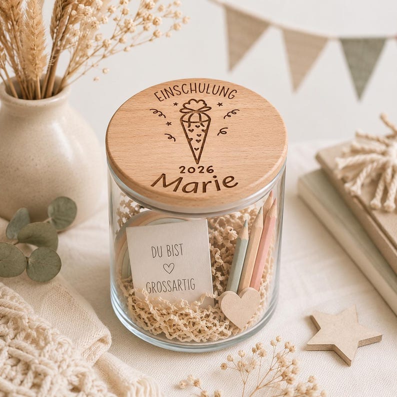 May include: A clear glass jar with a round wooden lid engraved with the text EINSCHULUNG 2026 Marie and a graphic of a cone. Inside the jar are pencils, a small heart-shaped wooden piece, and a card with the text DU BIST GROSSARTIG.