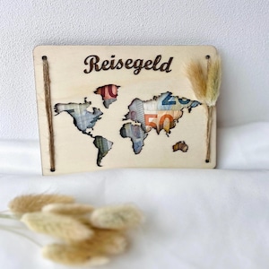 Könnte beinhalten: Holzschild mit Weltkarten-Design, gefüllt mit Währung und dem Wort "Reisegeld" in Schrift. Das Schild hat Schnurakzente und ein kleines Bündel getrockneter Blumen. Der Hintergrund ist weiß.