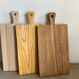 May include: Three rectangular wooden cutting boards with handles, each featuring a circular hole at the top. The boards display varying wood tones, ranging from a light pale beige to a warm golden yellow and a deeper brown grain pattern.