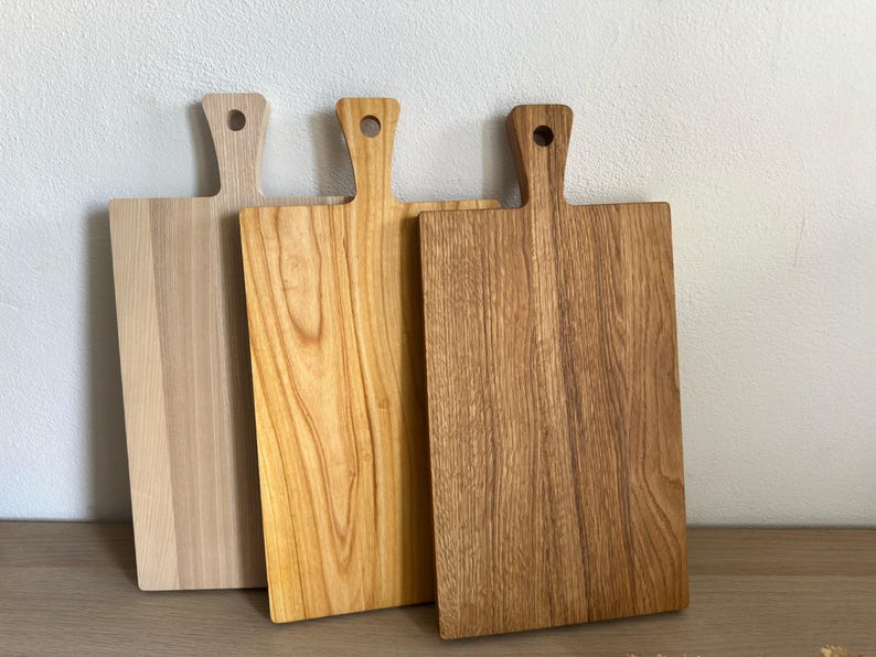 May include: Three rectangular wooden cutting boards with handles, each featuring a circular hole at the top. The boards display varying wood tones, ranging from a light pale beige to a warm golden yellow and a deeper brown grain pattern.