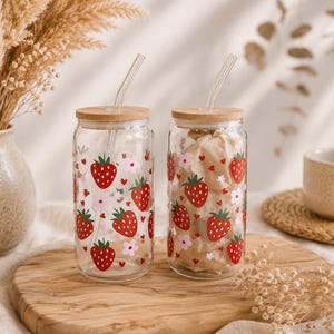 May include: Two clear glass tumblers with bamboo lids and glass straws, each decorated with a repeating pattern of red strawberries, small pink flowers, and tiny red hearts. The cylindrical glasses have a smooth finish and a wide opening.