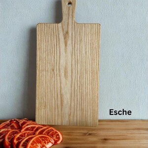 May include: A rectangular light-colored wooden cutting board with a handle and a circular hole at the top. The board features a prominent vertical wood grain pattern. The text "Esche" is printed in black on the lower right side.