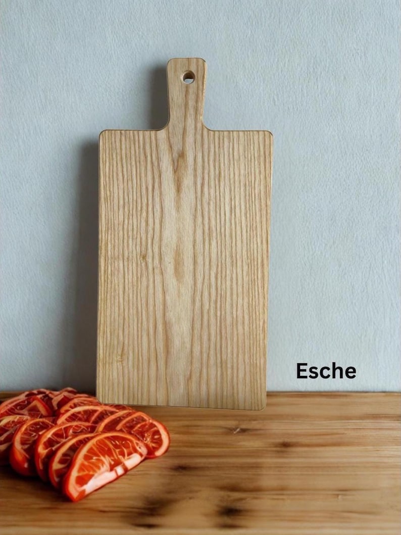 May include: A rectangular light-colored wooden cutting board with a handle and a circular hole at the top. The board features a prominent vertical wood grain pattern. The text "Esche" is printed in black on the lower right side.