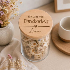 May include: A clear glass jar filled with small square items wrapped in floral paper, topped with a round wooden lid. The lid is engraved with the German text "Ein Glas voll Dankbarkeit" and the name "Liana" separated by a small heart icon.