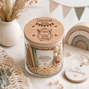 May include: A clear glass jar with a wooden lid engraved with a backpack, the text SCHULKIND 2026, and the name Clara. Inside are colored pencils, wooden beads, and a card reading Ein neues ABENTEUER beginnt. A round wooden tag reads Zur Einschulung.