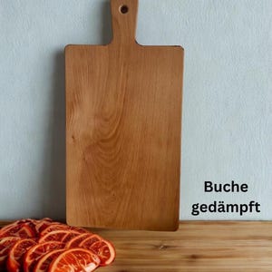 May include: A rectangular wooden cutting board with a handle and a circular hole at the top. The board has a light brown wood grain pattern. To the bottom left, there are several sliced orange citrus fruits. The text Buche ged&auml;mpft is visible.