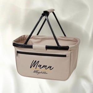Può includere: Borsa di tela beige per la spesa con manici neri e una cerniera nera. La borsa ha il testo "Mama Alltagsheldin" stampato in nero sul davanti.