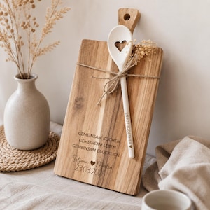 May include: A rectangular wooden cutting board with a handle and a white wooden spoon featuring a heart-shaped cutout. The board is engraved with the text GEMEINSAM KOCHEN GEMEINSAM LEBEN GEMEINSAM GL&Uuml;CKLICH, Tatjana, Merlin, and 23.03.2026. The spoon is tied with twine.