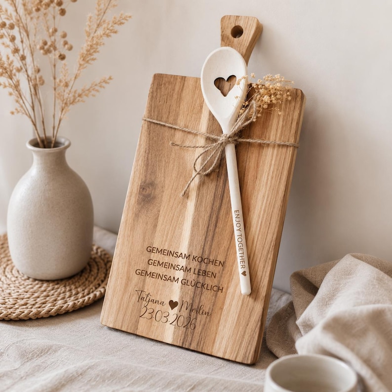May include: A rectangular wooden cutting board with a handle and a white wooden spoon featuring a heart-shaped cutout. The board is engraved with the text GEMEINSAM KOCHEN GEMEINSAM LEBEN GEMEINSAM GL&Uuml;CKLICH, Tatjana, Merlin, and 23.03.2026. The spoon is tied with twine.