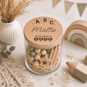 May include: A clear glass jar with a round wooden lid engraved with the letters A B C, the name Mattis, the word Schulkind, and the year 2026. The jar is filled with wooden beads, a star-shaped wooden cutout with a heart, and colored pencils.
