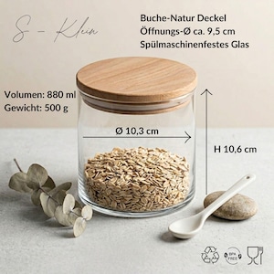 May include: A clear glass storage jar with a natural beech wood lid, filled with oats. Text indicates S-Klein, 880 ml volume, 500 g weight, 9.5 cm opening diameter, 10.3 cm diameter, and 10.6 cm height. Includes dishwasher-safe glass, BPA-free, and food-safe icons.