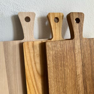 May include: Three rectangular wooden cutting boards with handles, each featuring a circular hole for hanging. The boards display varying natural wood grain patterns and tones, ranging from a light pale finish to a deeper, warm brown hue.