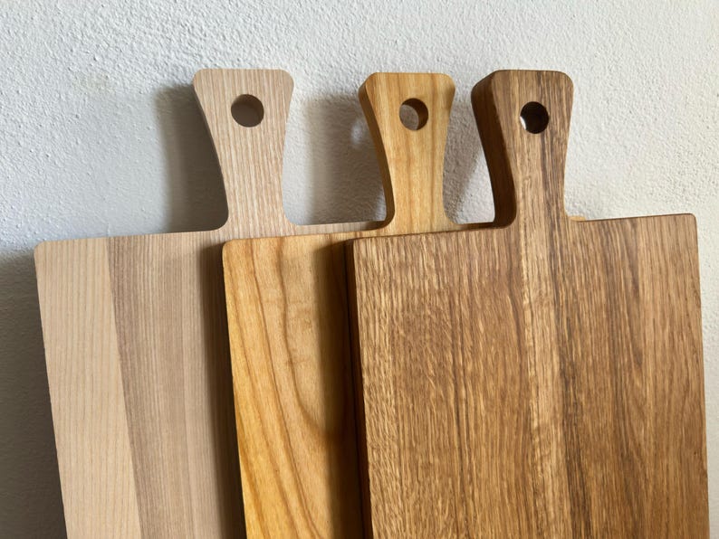 May include: Three rectangular wooden cutting boards with handles, each featuring a circular hole for hanging. The boards display varying natural wood grain patterns and tones, ranging from a light pale finish to a deeper, warm brown hue.