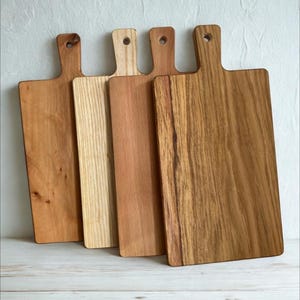 May include: Four rectangular wooden cutting boards with handles, each featuring a distinct wood grain pattern and varying shades of brown, ranging from light tan to a deep, rich amber, all aligned side by side in a vertical orientation.