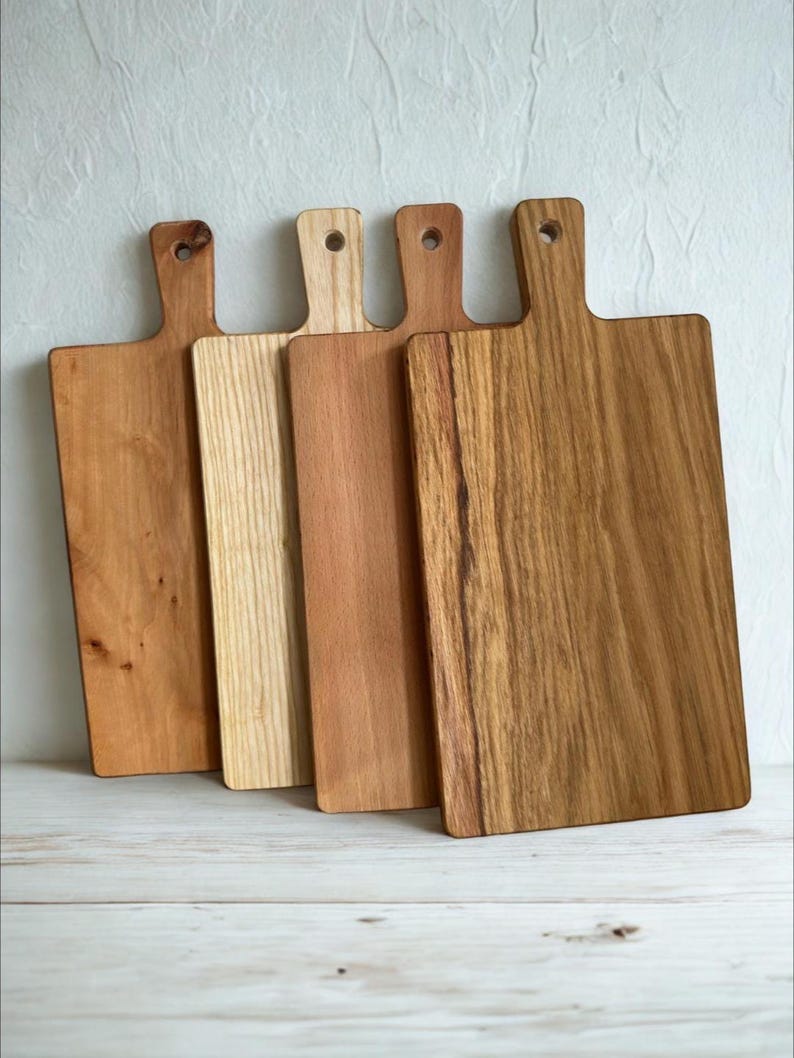 May include: Four rectangular wooden cutting boards with handles, each featuring a distinct wood grain pattern and varying shades of brown, ranging from light tan to a deep, rich amber, all aligned side by side in a vertical orientation.