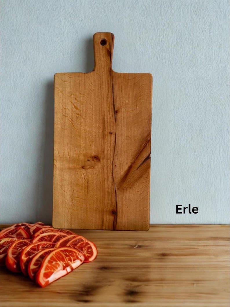 May include: A rectangular wooden cutting board with a handle and a hole for hanging, featuring a natural light brown wood grain pattern. The word Erle is printed in black text to the right of the board, near sliced orange fruit.