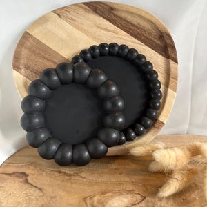 Könnte beinhalten: Zwei schwarze Untersetzer mit Perlen liegen auf einem Holzserviertablett mit Streifenmuster. Die Untersetzer haben eine runde Form mit einem erhabenen, perlenbesetzten Rand. Das Tablett ist aus hellem und dunklem Holz.