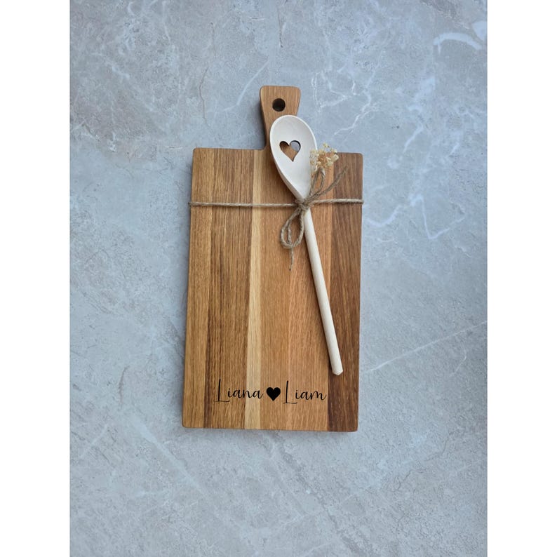 May include: A rectangular wooden cutting board with a handle and a hole, featuring the engraved text Liana heart Liam. A light-colored wooden spoon with a heart-shaped cutout in the bowl is tied to the board with twine.