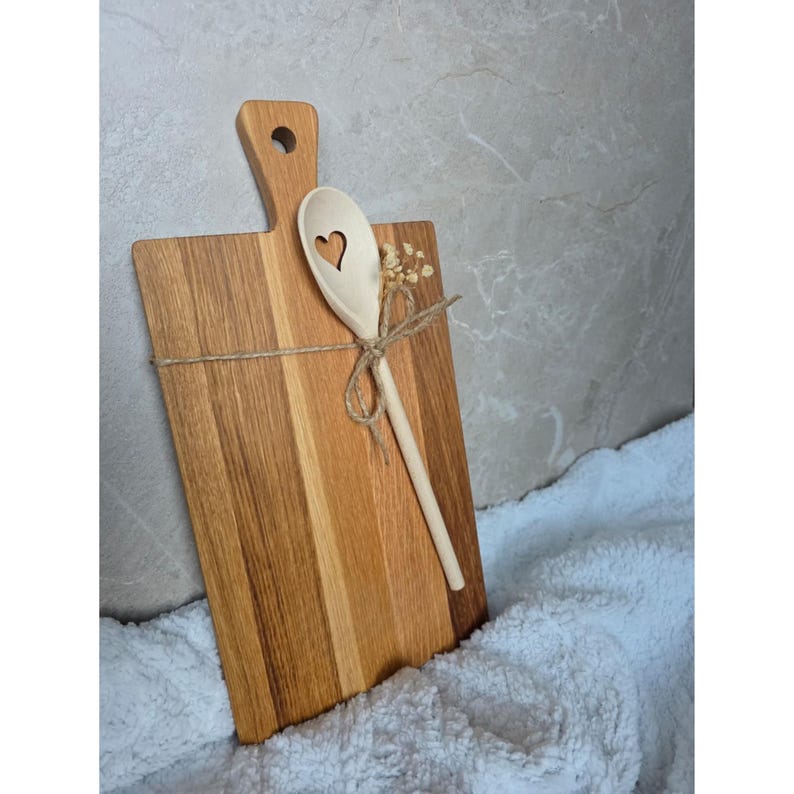 May include: A rectangular wooden cutting board with a handle featuring a circular hole, paired with a light-colored wooden spoon that has a heart-shaped cutout in the bowl. The spoon is tied to the board with twine and a small dried flower.