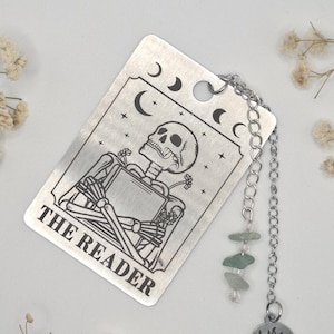 Könnte beinhalten: Silberner Metall-Lesezeichen mit einer Skelett-Illustration von einer Tarotkarte und dem Text "THE READER". Das Lesezeichen hat eine Kette mit grünen Perlen und einem Sonnenanhänger. Das Design umfasst Halbmonde und Sterne.