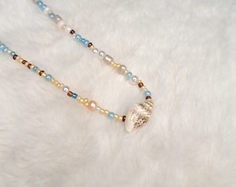 Puede incluir: Un collar delicado hecho con pequeñas cuentas blancas, marrones y azules, acentuado con un colgante de concha.