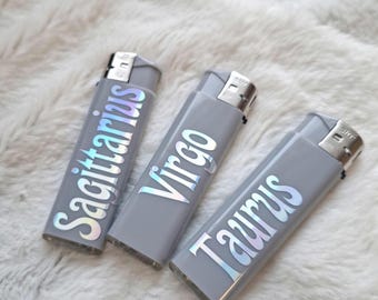 Puede incluir: Tres encendedores grises con texto holográfico de los signos del zodiaco: Sagitario, Virgo y Tauro. Los encendedores están colocados sobre una superficie suave y blanca.