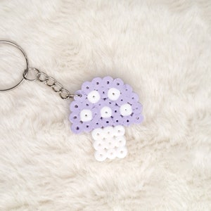 Puede incluir: Un llavero en forma de hongo morado y blanco hecho de cuentas perler. El llavero est&aacute; unido a una cadena plateada.