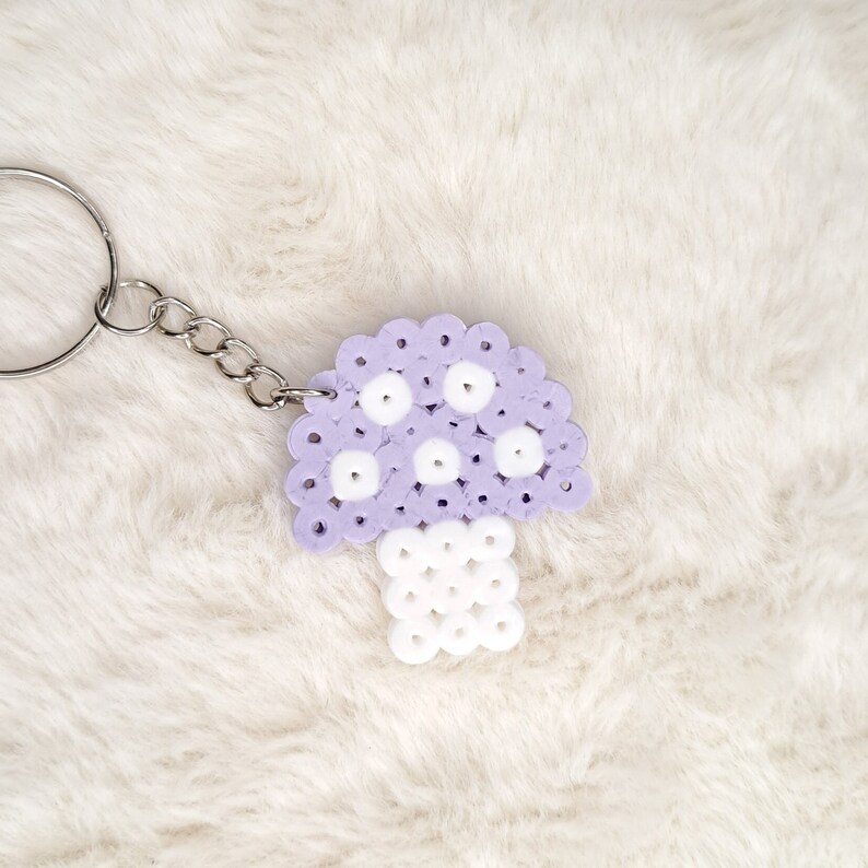 Puede incluir: Un llavero en forma de hongo morado y blanco hecho de cuentas perler. El llavero est&aacute; unido a una cadena plateada.