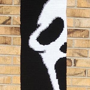May include: A rectangular crochet wall hanging featuring a black background with a large, white, stylised ghost face design. The piece is suspended from a wooden dowel with a black string attached for hanging, creating a high-contrast graphic display.