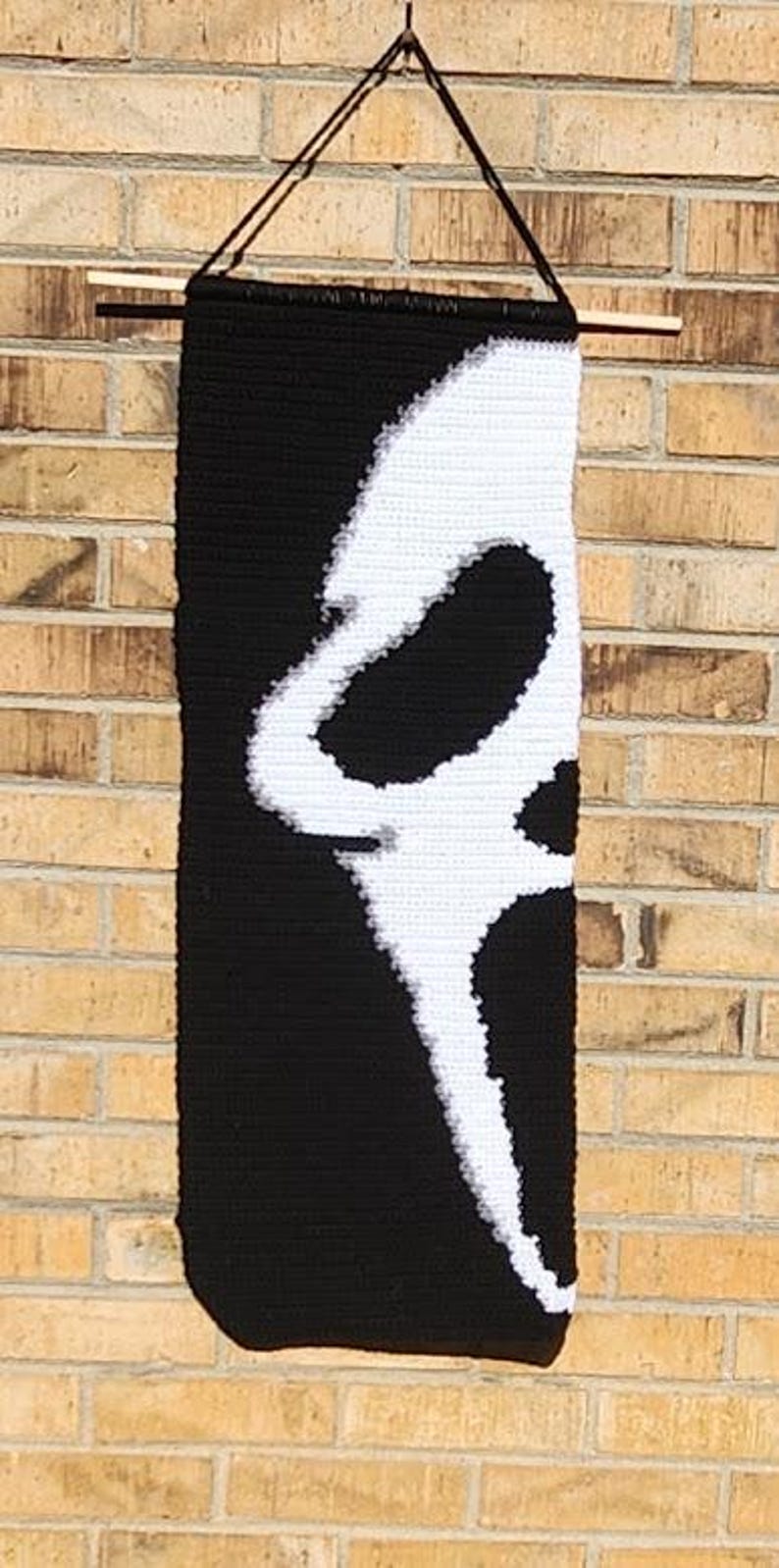 May include: A rectangular crochet wall hanging featuring a black background with a large, white, stylised ghost face design. The piece is suspended from a wooden dowel with a black string attached for hanging, creating a high-contrast graphic display.