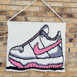 May include: A rectangular crochet wall hanging featuring a pixelated graphic of a sneaker in white, grey, black, and pink yarn. The textile art is suspended from a wooden dowel with a white crocheted string for hanging, showcasing detailed stitching.