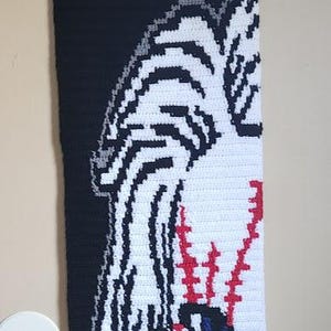 May include: A rectangular crochet wall hanging featuring a black background with a white, red, and blue graphic design of a character face. The textile is suspended from a wooden dowel with a black yarn cord for hanging, showcasing detailed stitch work.