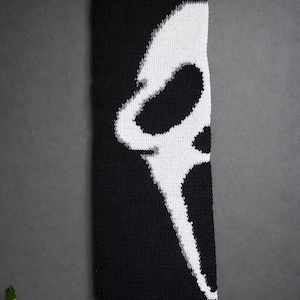 Puede incluir: Tapiz de pared tejido en blanco y negro con el diseño de la máscara de Scream. La pieza rectangular está suspendida de una clavija de madera con un cordón negro. La máscara es blanca sobre un fondo negro.