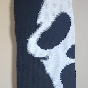 May include: A rectangular crochet wall hanging featuring a high-contrast black and white design of a stylised ghost mask. The piece is suspended from a wooden dowel with a black yarn cord, showcasing intricate stitch work and a monochromatic aesthetic.