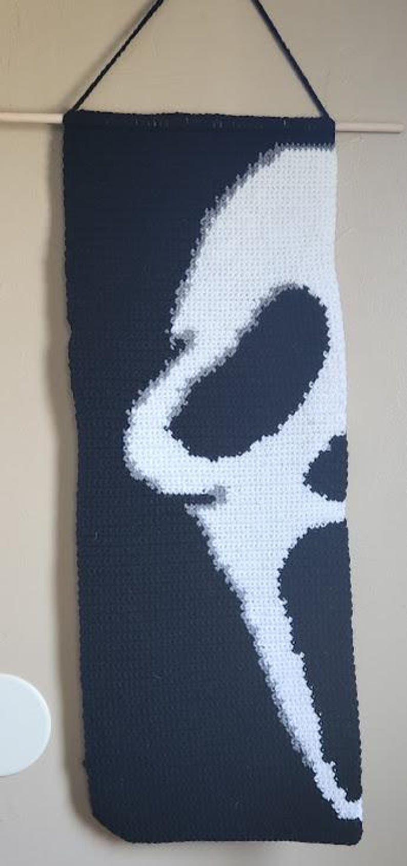 May include: A rectangular crochet wall hanging featuring a high-contrast black and white design of a stylised ghost mask. The piece is suspended from a wooden dowel with a black yarn cord, showcasing intricate stitch work and a monochromatic aesthetic.