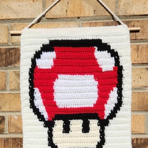 May include: A rectangular crochet wall hanging featuring a pixelated red, white, and black mushroom design. The piece is suspended from a wooden dowel with a white yarn string, showcasing a detailed stitch pattern against a cream-colored background.