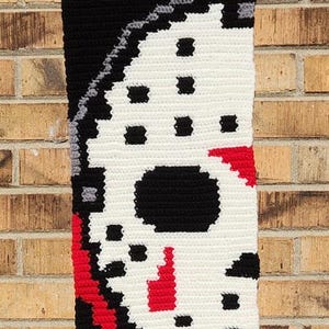 May include: A rectangular crochet wall hanging featuring a pixelated white hockey mask design on a black background. The mask includes black eye and breathing holes, red triangular accents, and a red drip pattern at the bottom, suspended by a wooden dowel.