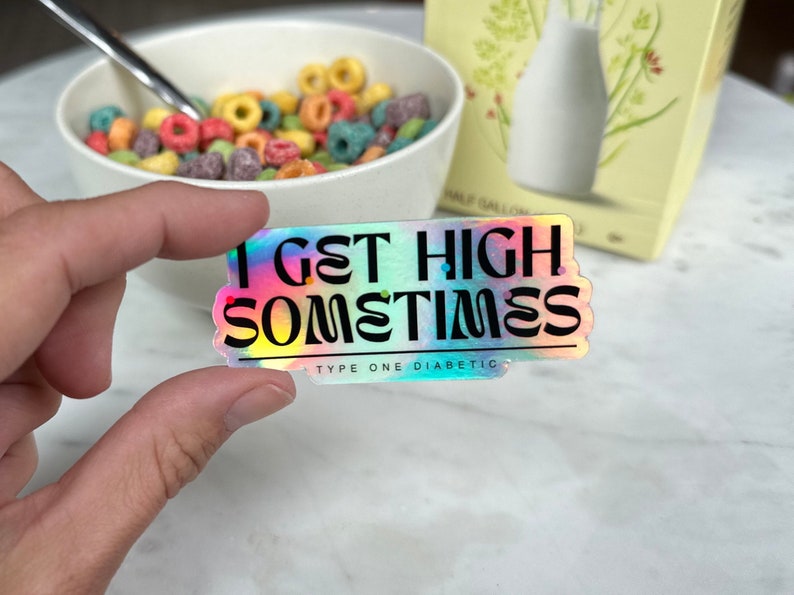Op de afbeelding: Een holografische sticker met een regenboogachtergrond en zwarte tekst die "I get high sometimes" en "Type One Diabetic" zegt.