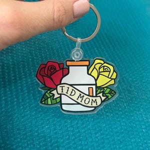 Op de afbeelding: Een transparante acryl sleutelhanger met een cartoonillustratie van een pillenfles met de tekst "TID MOM" op een banner. De fles wordt omringd door twee rozen, een rode en een gele.